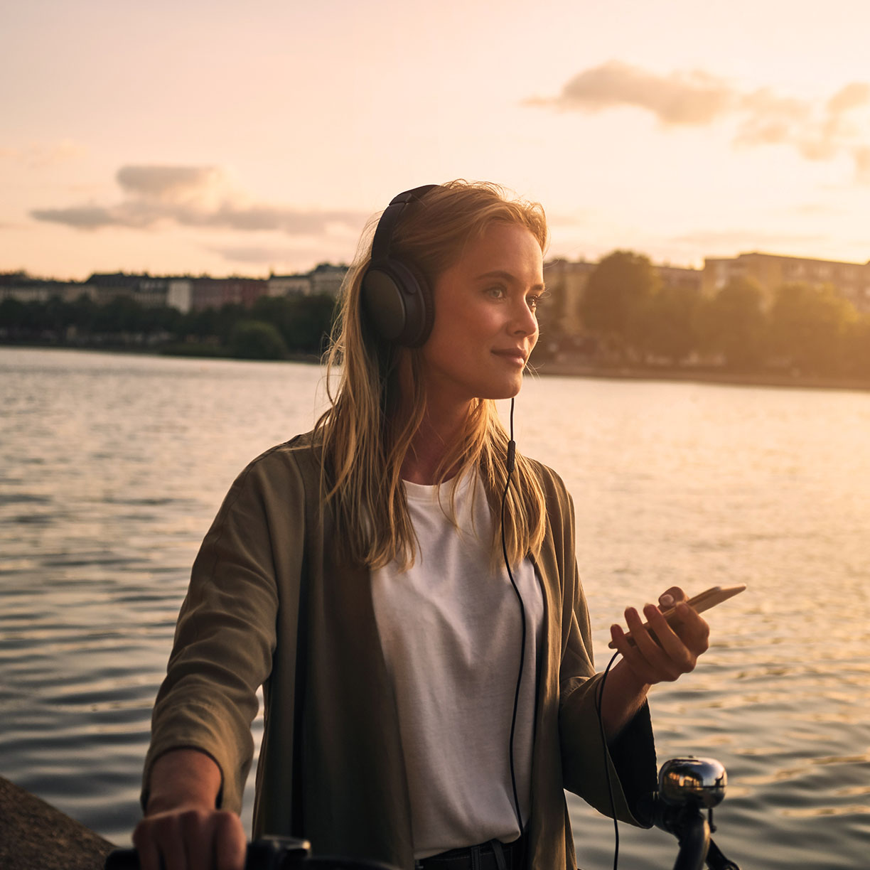 Kvinde med hovedtelefoner står ved vandet i aftensol og holder sin mobiltelefon
