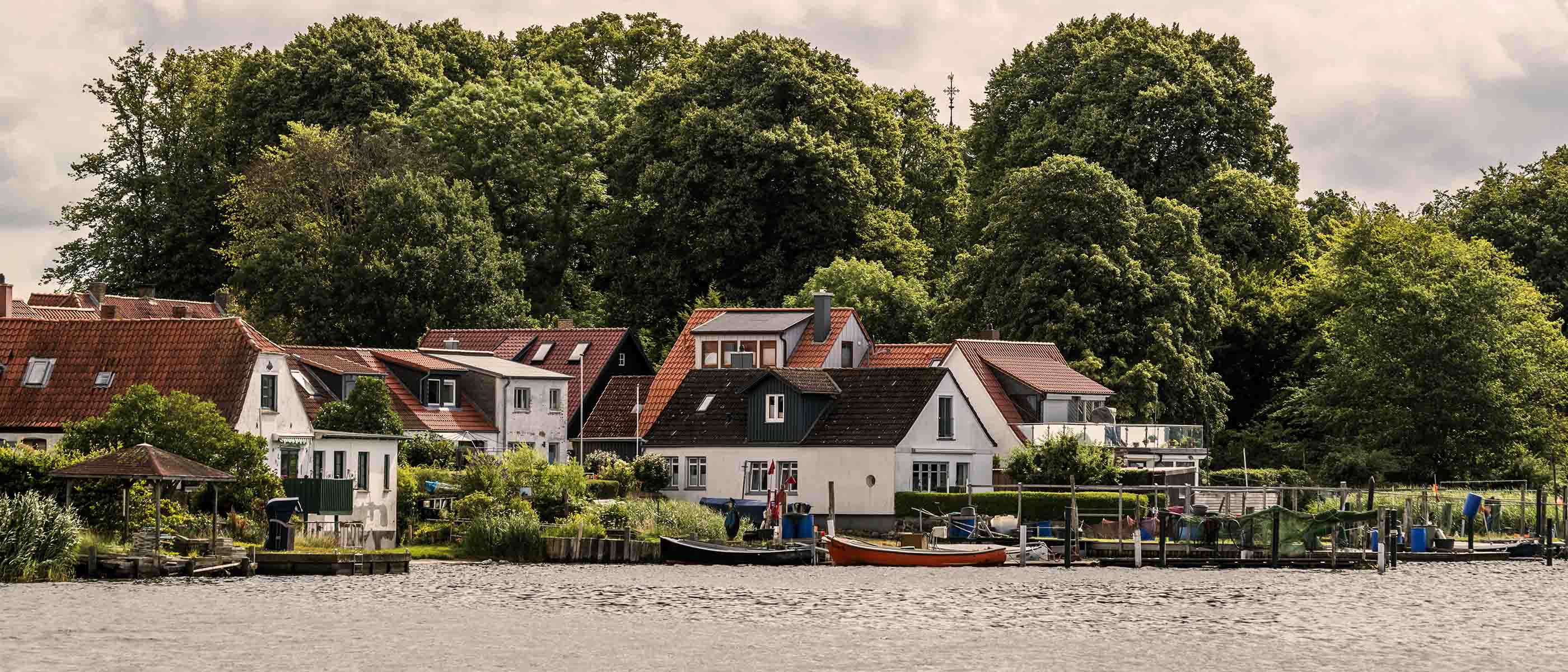Landsby ved vand med små huse, både og grønne træer i baggrunden
